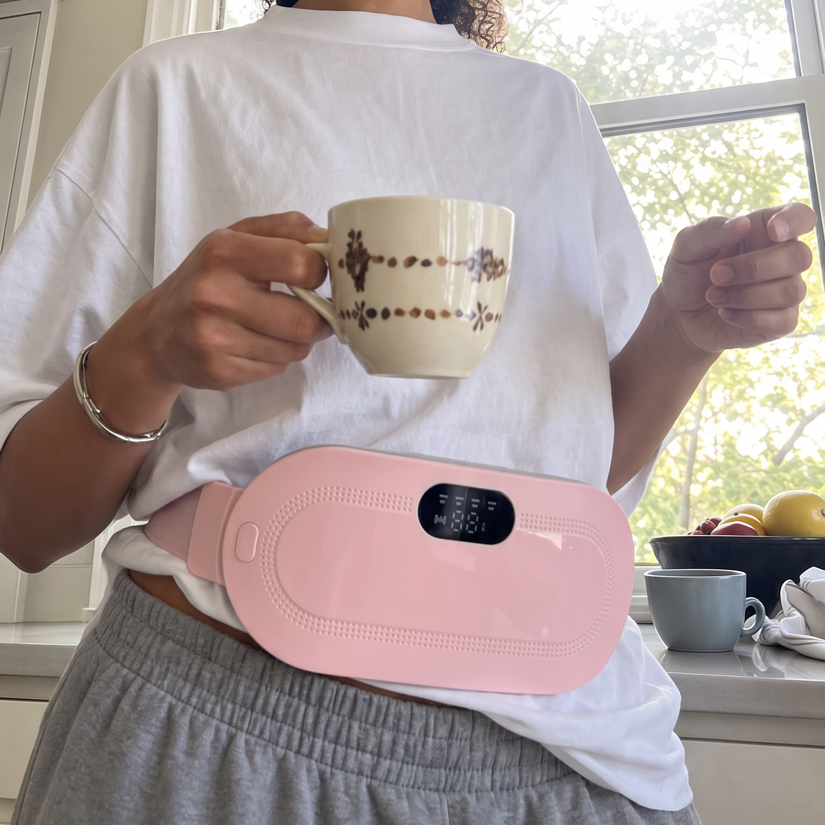 A woman in a white t-shirt and gray sweatpants holds a patterned mug while wearing a pink waist heating pad around her stomach in a bright kitchen setting.